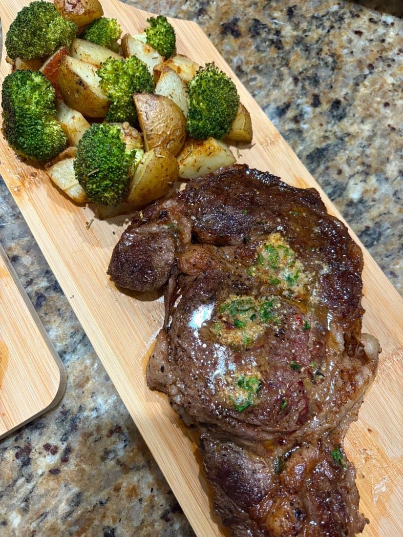 Pan-seared ribeye topped with herb butter, served with roasted potatoes and broccoli on a wooden board